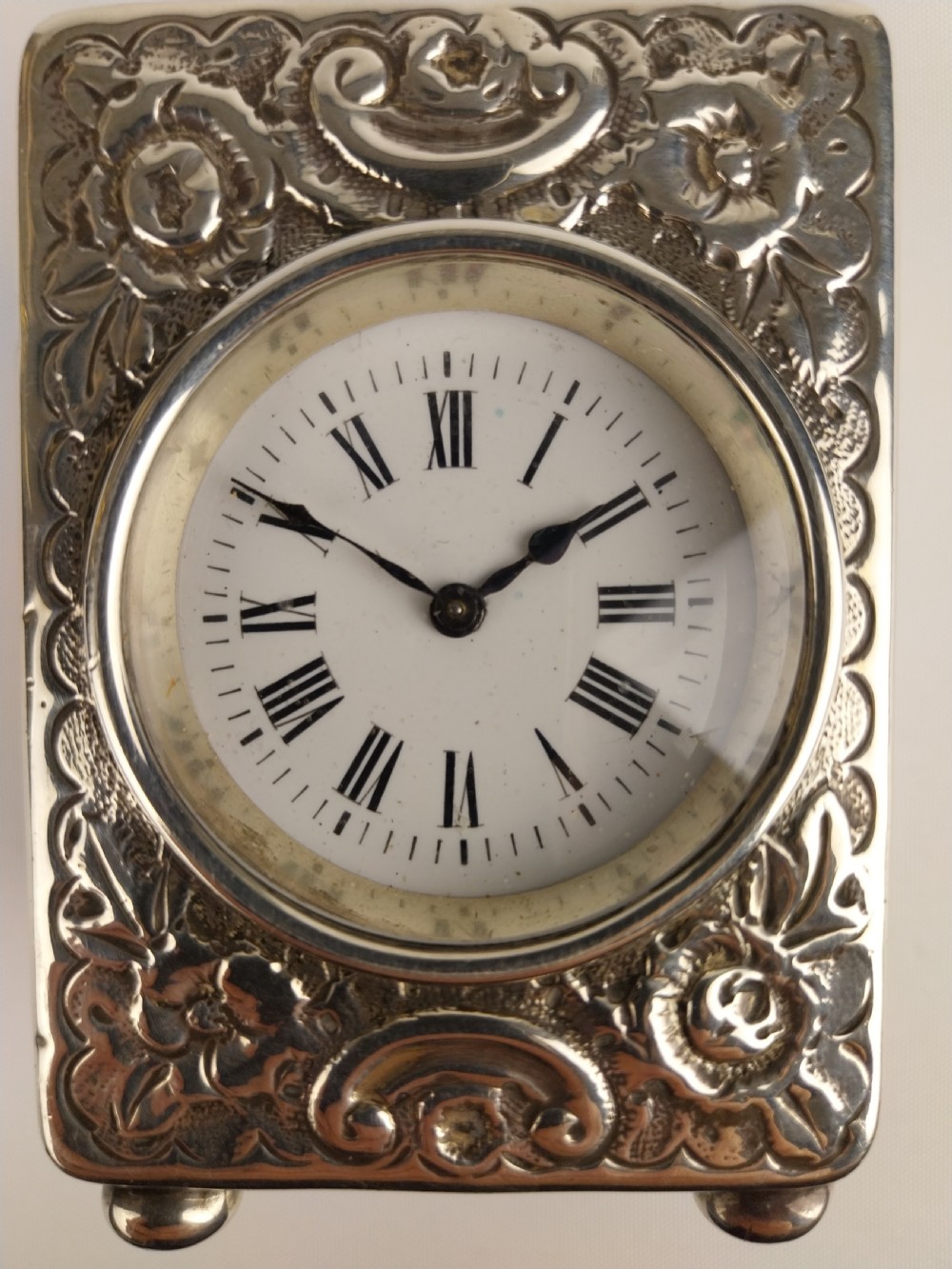 A rectangular miniature silver carriage clock featuring floral relief chasing, a curved carrying handle, ball feet, and a white enamel dial with Roman numerals. View XIV