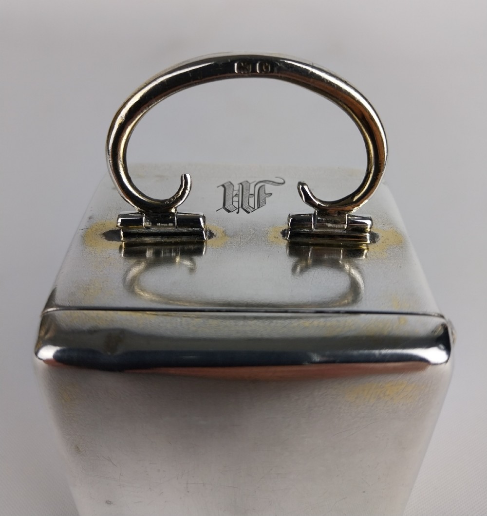 A miniature rectangular silver carriage clock with a curved top loop handle, bun feet, and a white circular dial featuring black Roman numerals. View XI