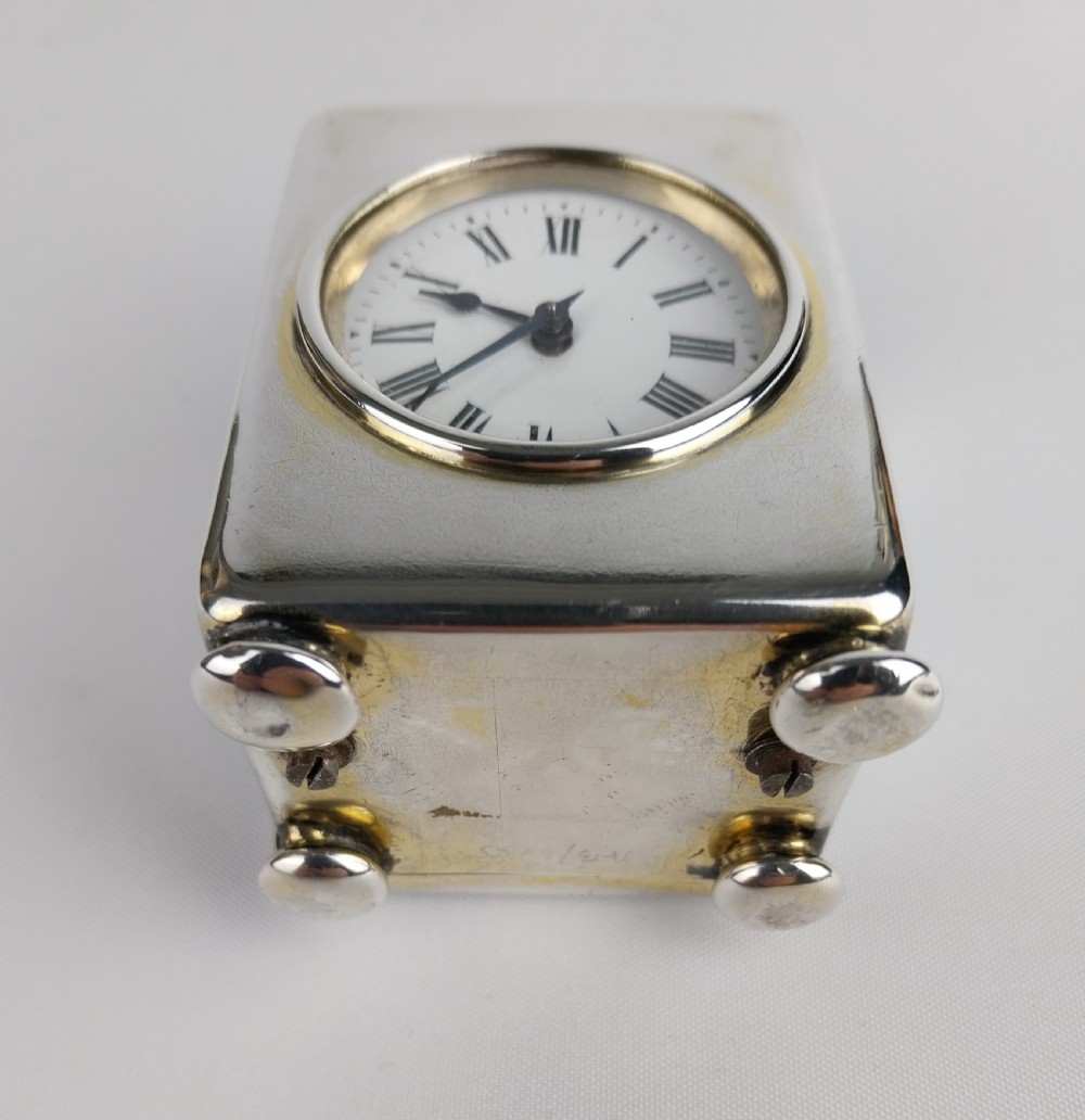 A miniature rectangular silver carriage clock with a curved top loop handle, bun feet, and a white circular dial featuring black Roman numerals. View XIV