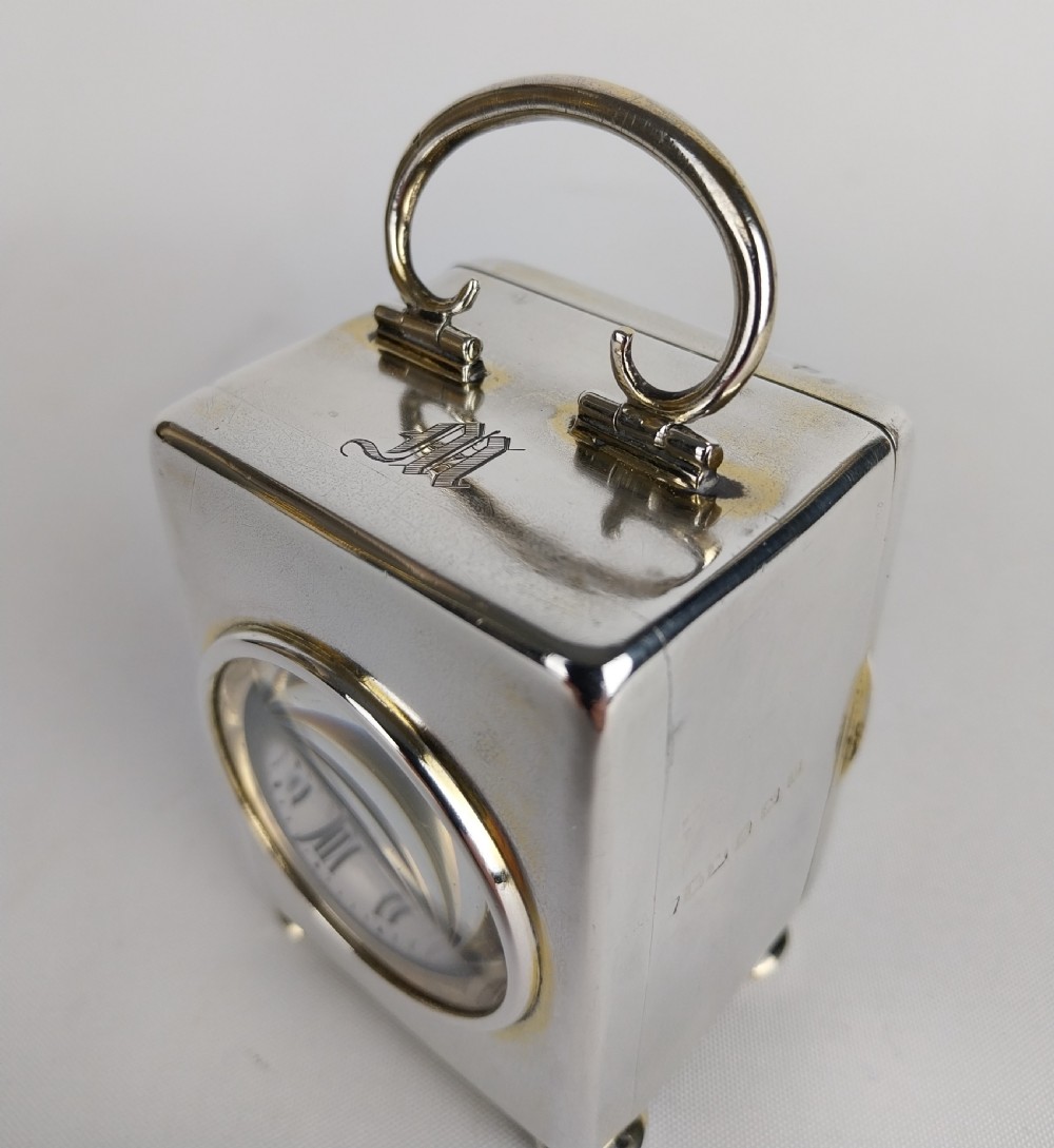 A miniature rectangular silver carriage clock with a curved top loop handle, bun feet, and a white circular dial featuring black Roman numerals. View XVIII