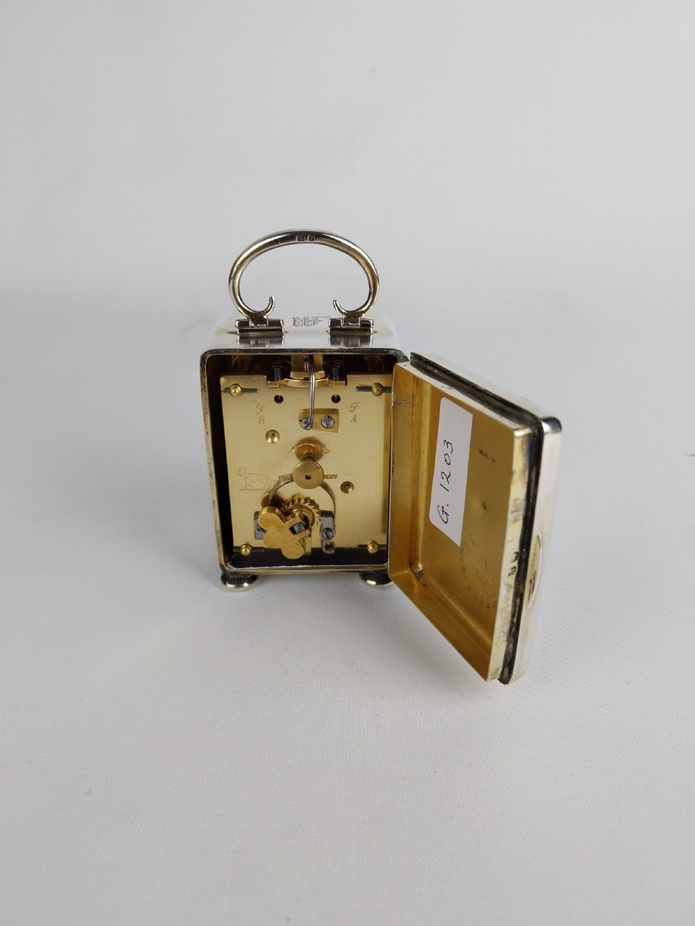 A miniature rectangular silver carriage clock with a curved top loop handle, bun feet, and a white circular dial featuring black Roman numerals. View XIX