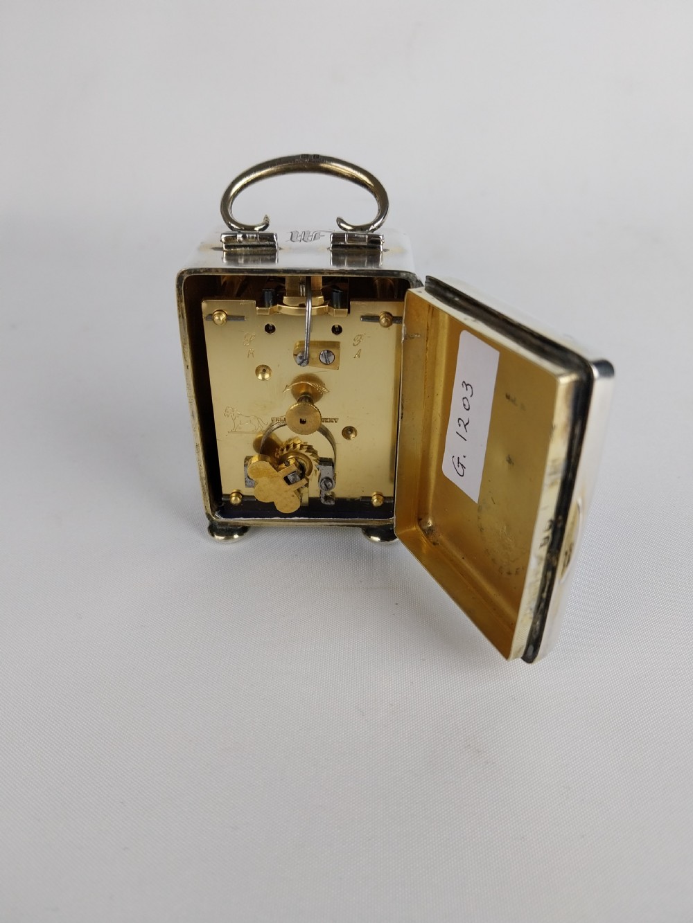 A miniature rectangular silver carriage clock with a curved top loop handle, bun feet, and a white circular dial featuring black Roman numerals. View XX