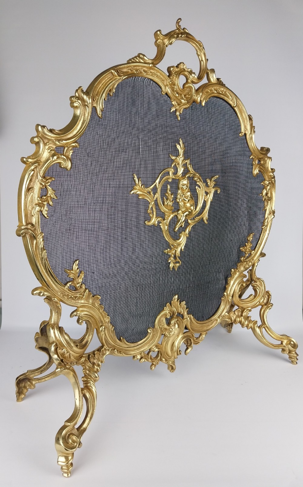 A French Louis XV style gilt bronze fire screen featuring an asymmetrical rocaille frame, central cherub motif, and woven metal mesh. View X