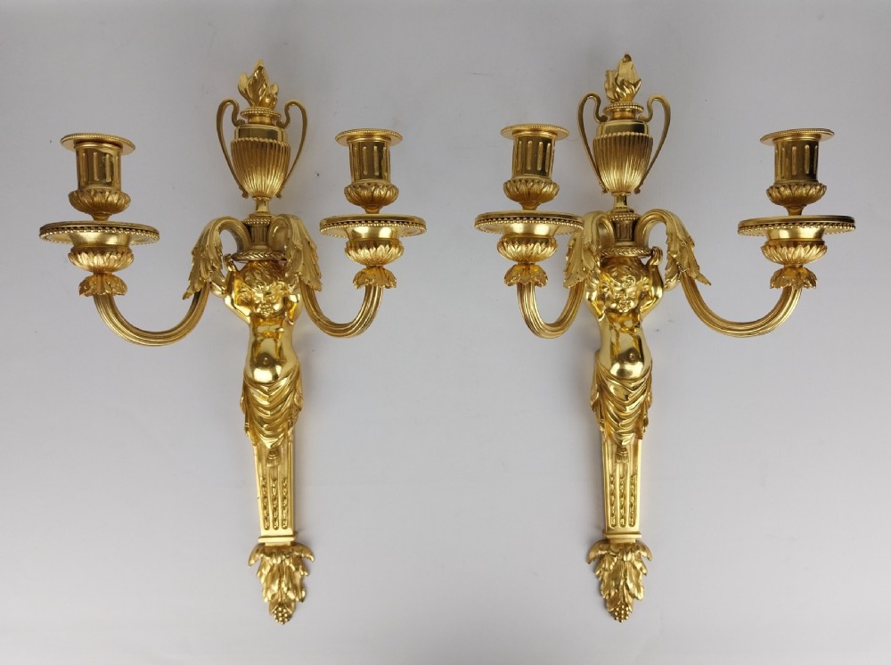 A pair of French Louis XVI style gilt bronze wall sconces featuring central cherub figures holding two curving candle arms topped with urn finials.