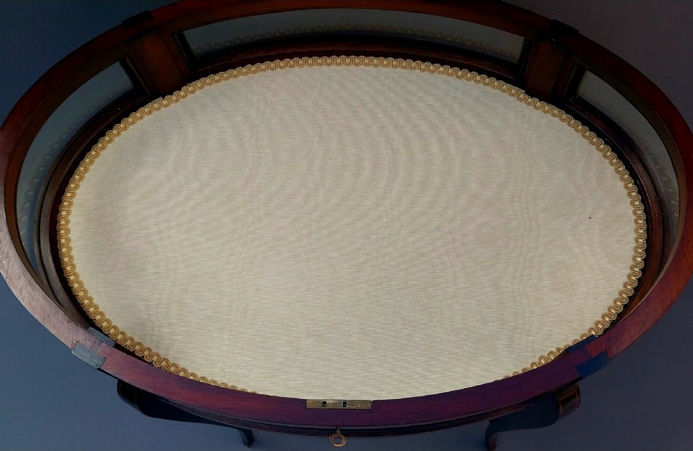 An oval Edwardian mahogany bijouterie table featuring glass side panels, a glazed top, satinwood inlaid rim, and slender cabriole legs joined by a curved stretcher. View XVI