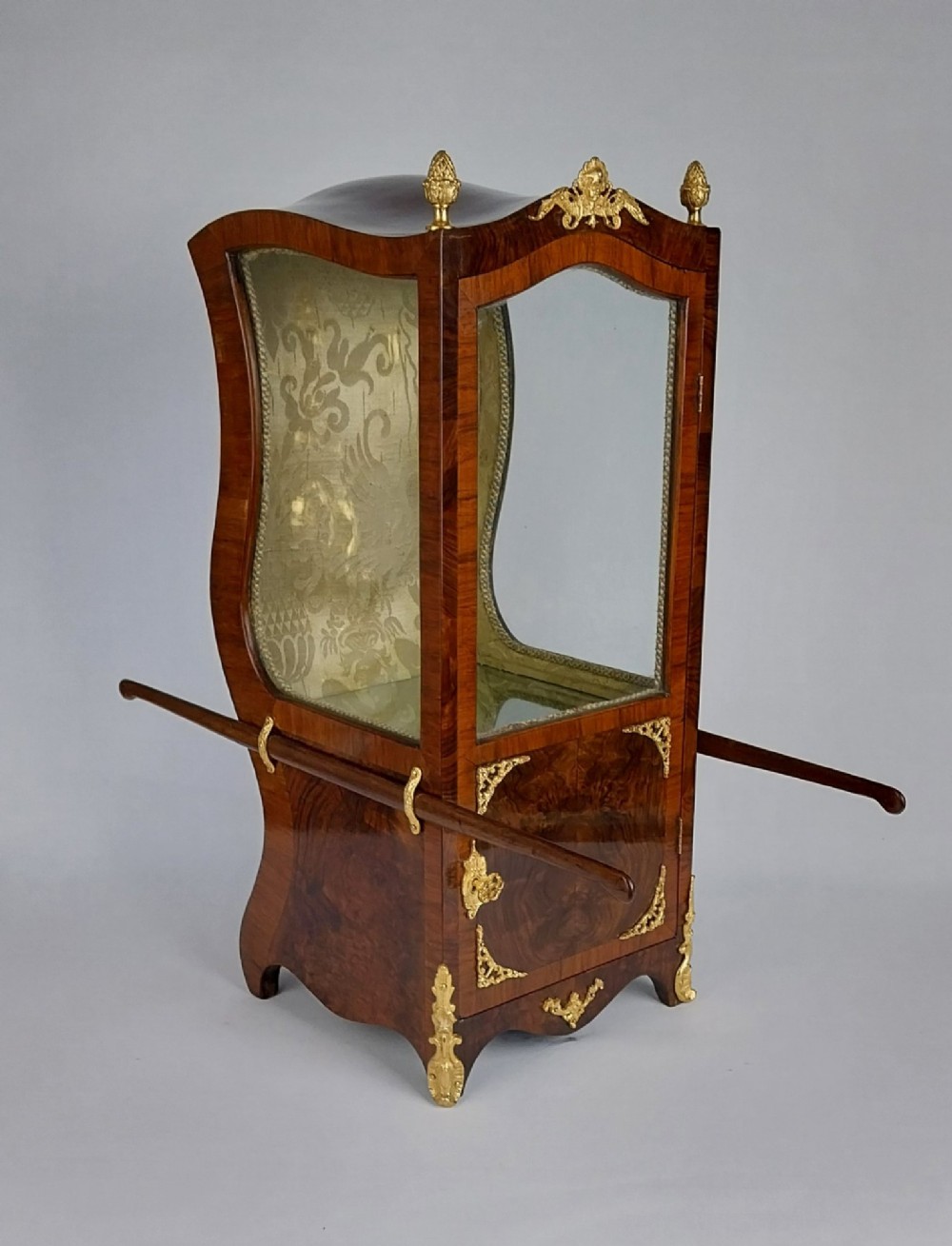 A French tabletop sedan chair vitrine cabinet crafted from polished brown wood veneer with gilt-bronze mounts, glazed side panels, removable carrying poles, and a silk damask interior.