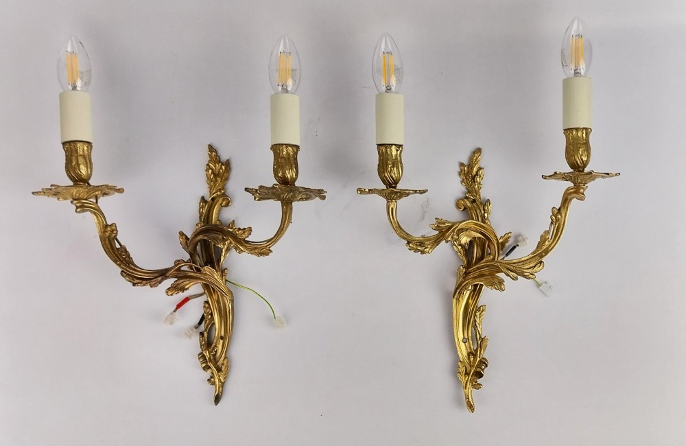 A pair of French Rococo style gilt bronze two-arm wall sconces featuring scrolling acanthus leaf backplates and candle-style sockets.