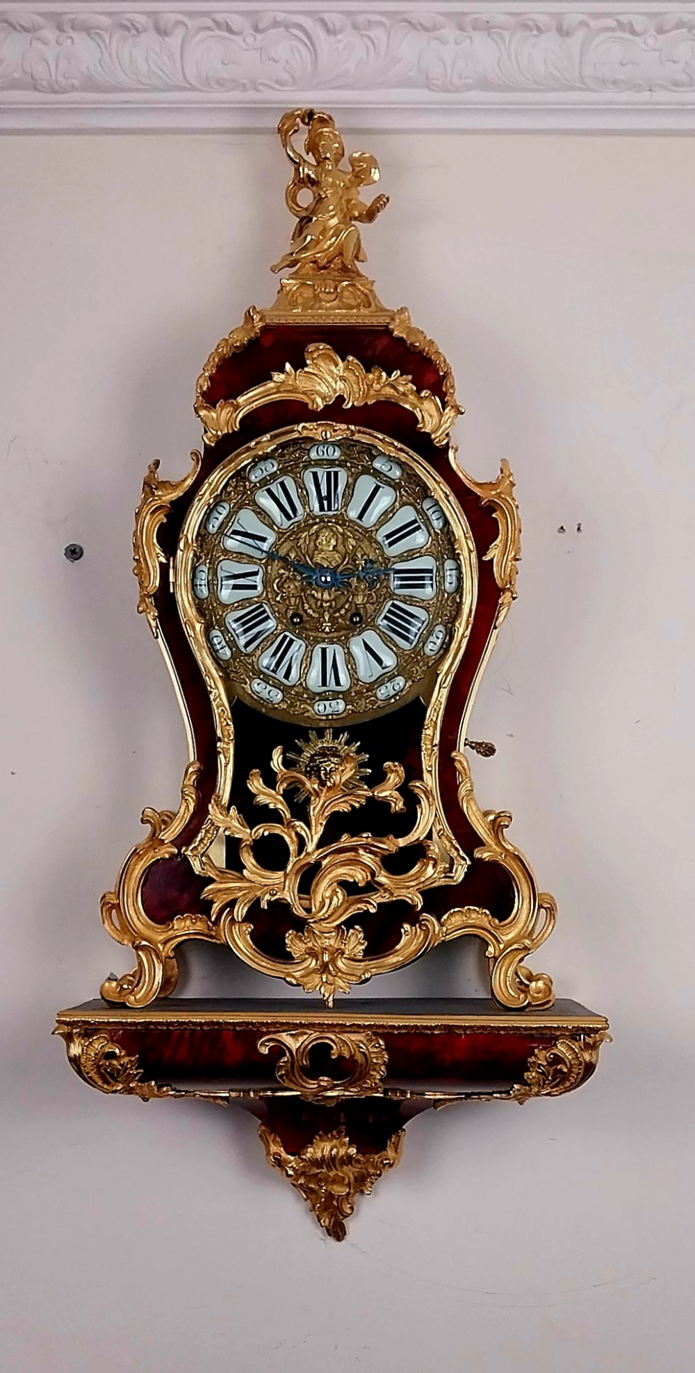 An antique French Rococo revival Boulle wall clock featuring a red tortoiseshell case adorned with elaborate gilt bronze mounts, a figure finial, and an enamel cartouche dial on a matching wall bracket.
