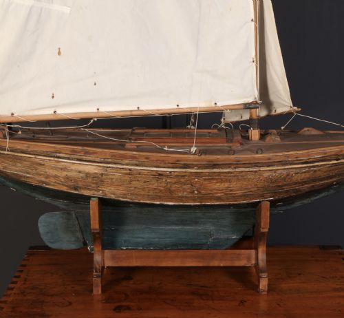 fig 1. An antique wooden pond yacht with off-white fabric sails, a carved brown hull with a teal painted lower section, mounted on a wooden display stand.