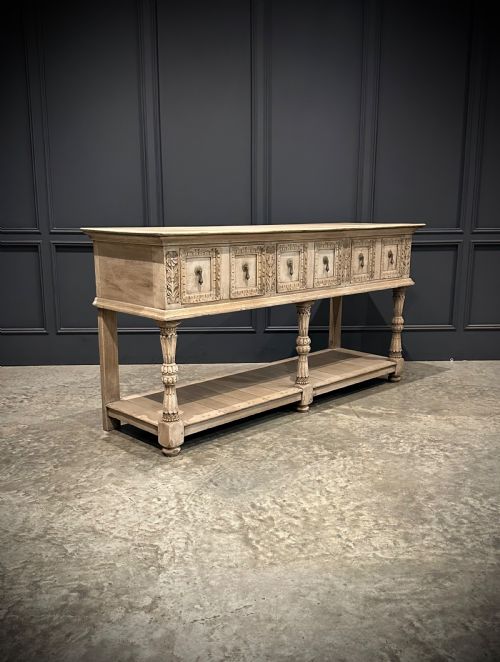 fig 1. A long carved bleached oak potboard dresser base featuring relief carved drawer fronts, turned support legs, brass drop handles, and an open lower display shelf.