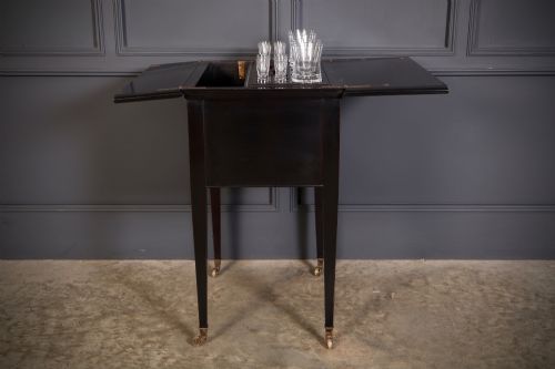 fig 1. An ebonised wooden mechanical drinks table featuring opening top leaves that elevate a tray set with clear glassware, resting on tapered legs with brass castors.