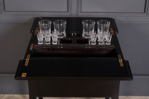 fig 2. An ebonised wooden mechanical drinks table featuring opening top leaves that elevate a tray set with clear glassware, resting on tapered legs with brass castors.