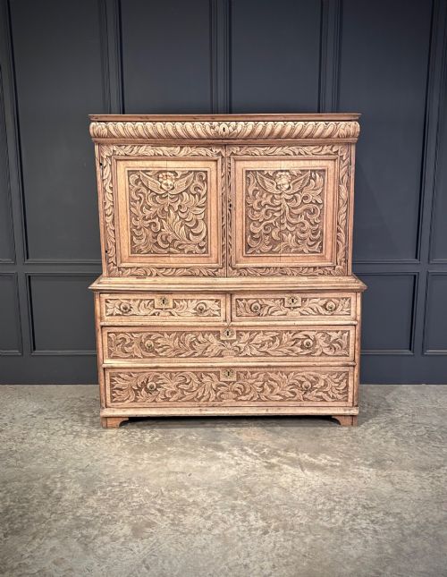 fig 1. An 18th-century light oak two-piece cupboard on chest featuring detailed hand-carved foliate panels, cherub motifs, and carved drawer fronts.