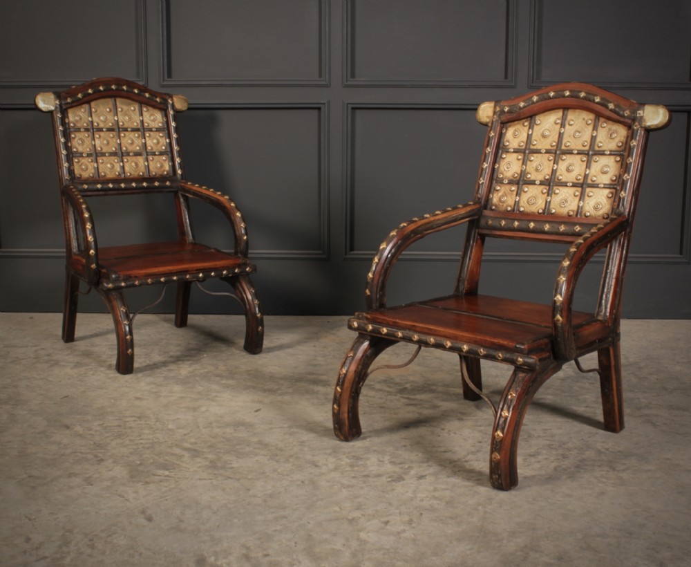 A pair of dark hardwood low armchairs featuring decorative brass studs, embossed brass backrest panels, curved front legs, and metal corner accents.