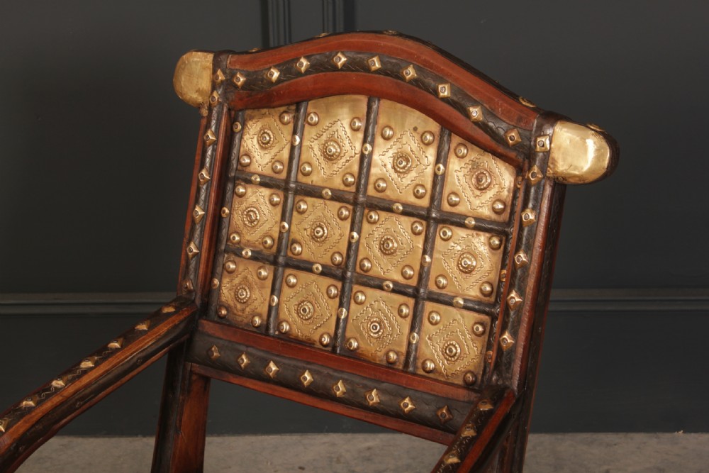 A pair of dark hardwood low armchairs featuring decorative brass studs, embossed brass backrest panels, curved front legs, and metal corner accents. View VI