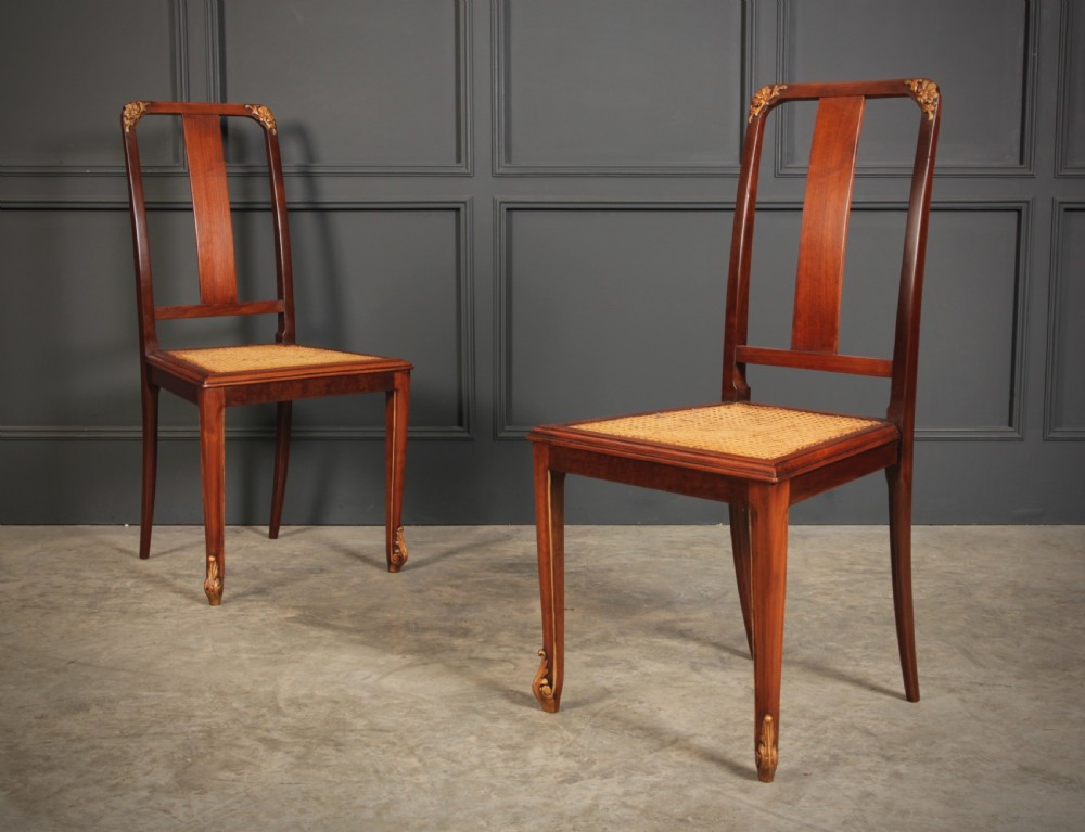 A pair of polished walnut bedroom chairs with woven cane seats, central splat backs, and carved gilt accents at the top corners and feet.