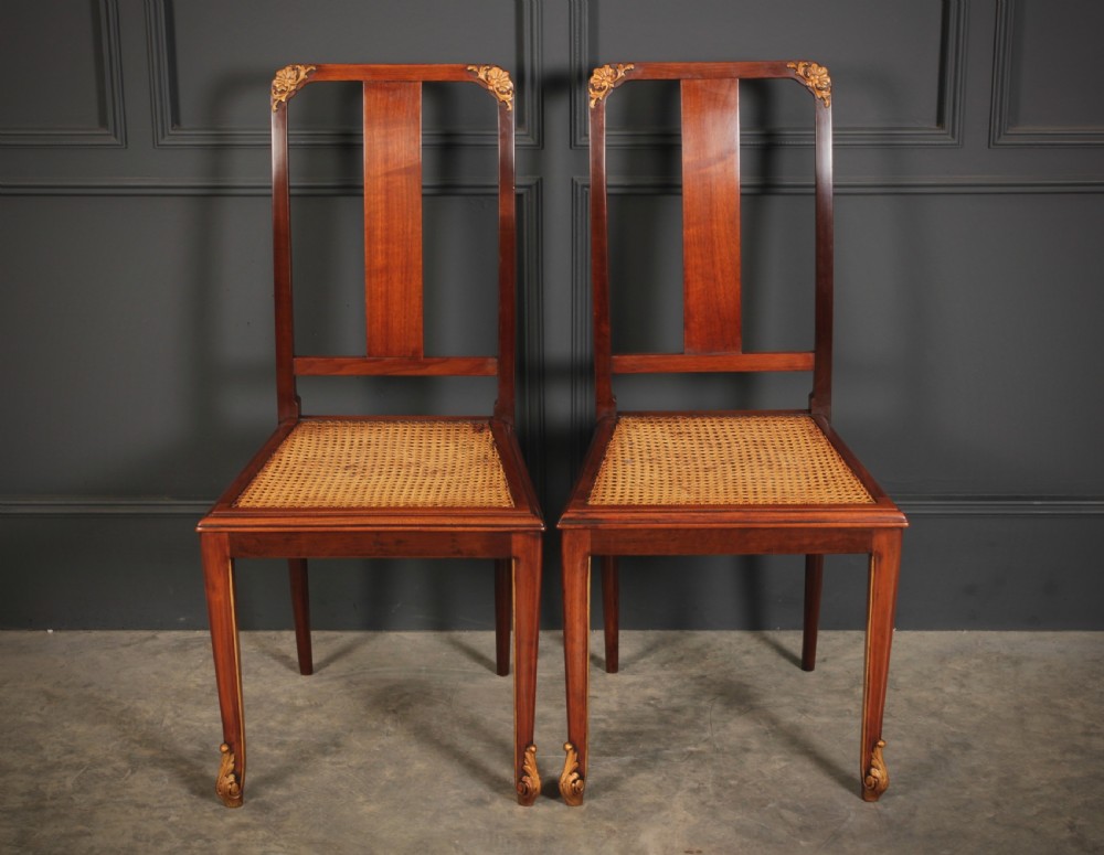 A pair of polished walnut bedroom chairs with woven cane seats, central splat backs, and carved gilt accents at the top corners and feet. View VII