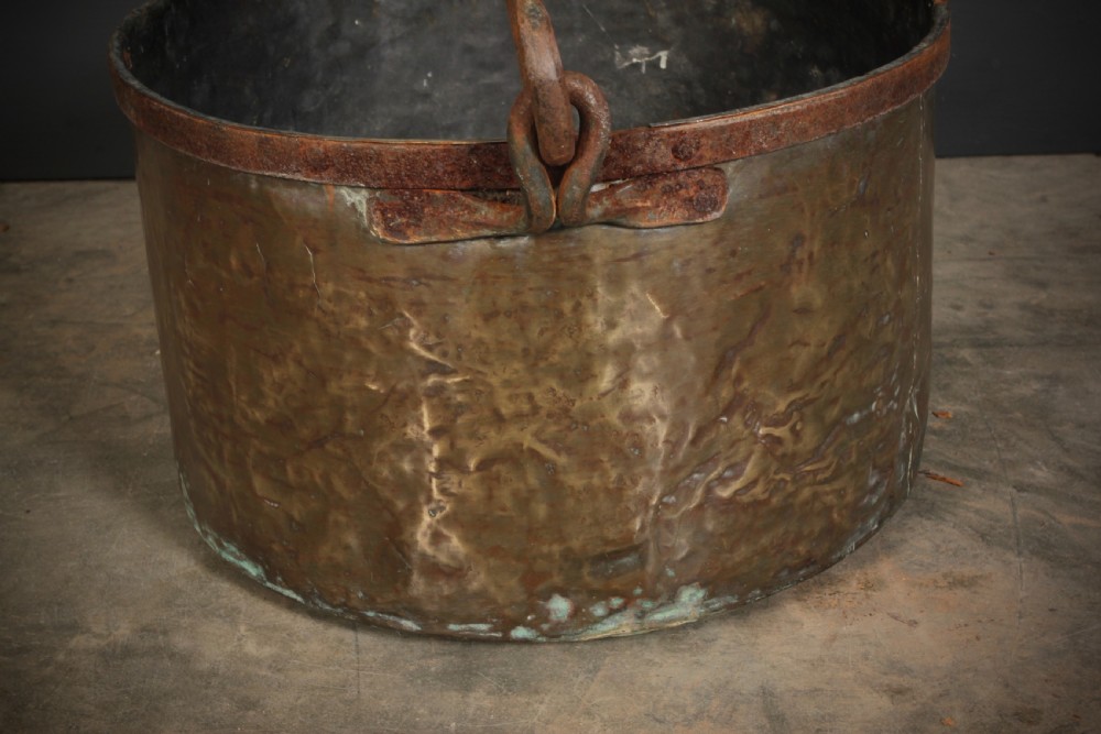 A large, hand-hammered Victorian brass cauldron featuring an iron rim collar and a heavy wrought iron swing handle. View VI
