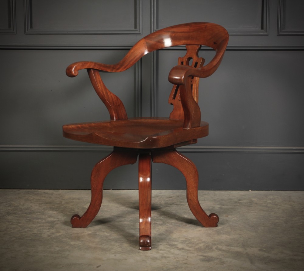 A Victorian mahogany swivel desk chair featuring a curved backrest with a carved central splat, shaped armrests, a solid saddled seat, and four splayed cabriole legs. View VI
