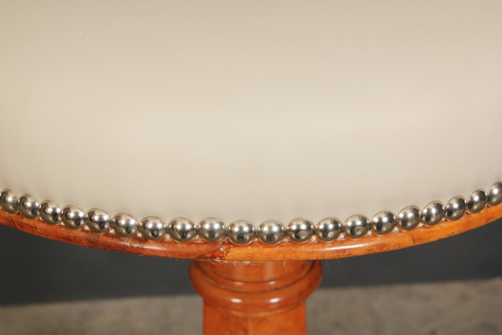 An Art Deco birds eye maple music stool featuring a round cream upholstered swivel seat with brass stud trimming above a faceted pedestal and octagonal base. View VII