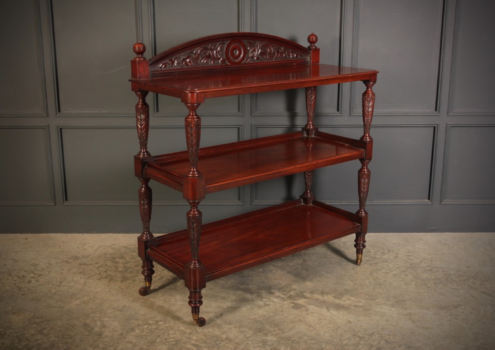 A Victorian mahogany three-tier buffet featuring carved turned column supports, a scrolled decorative backboard, and brass castors.