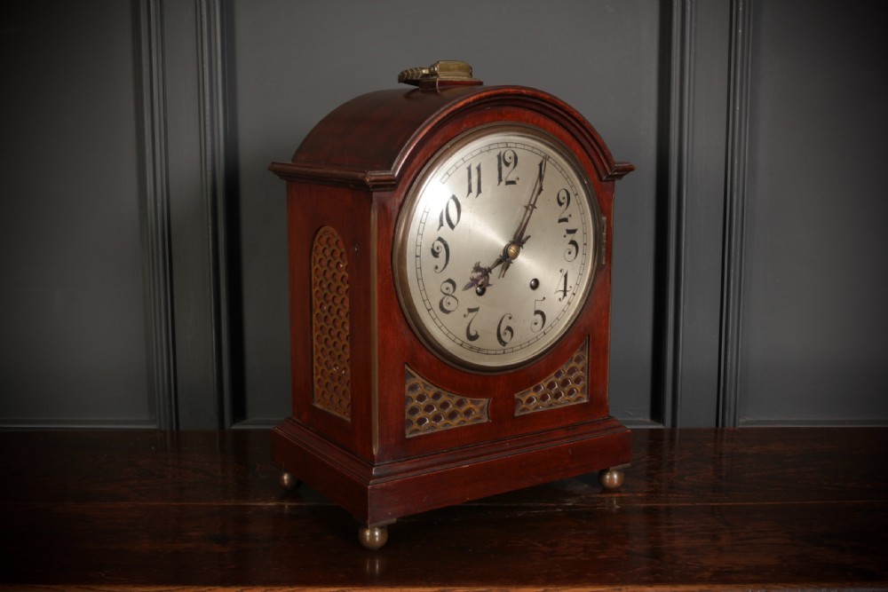 An antique arched mahogany bracket clock featuring brass fretwork panels, a circular dial with Arabic numerals, and small brass ball feet.
