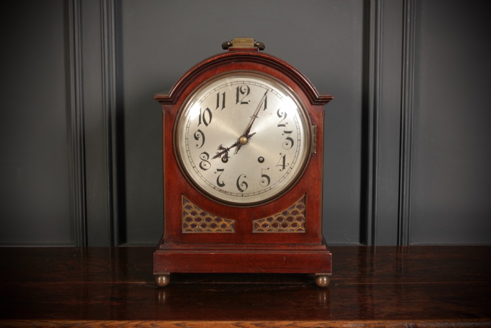An antique arched mahogany bracket clock featuring brass fretwork panels, a circular dial with Arabic numerals, and small brass ball feet. View II
