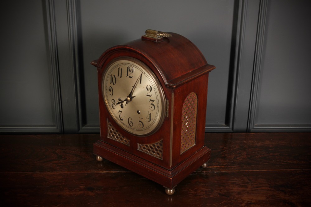 An antique arched mahogany bracket clock featuring brass fretwork panels, a circular dial with Arabic numerals, and small brass ball feet. View III
