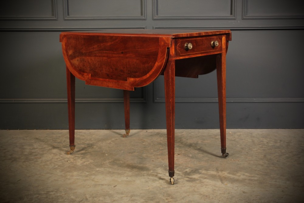 A Georgian mahogany Pembroke table featuring two drop leaves, subtle line inlay, a single front drawer with round brass handles, and tapered square legs on brass castors.