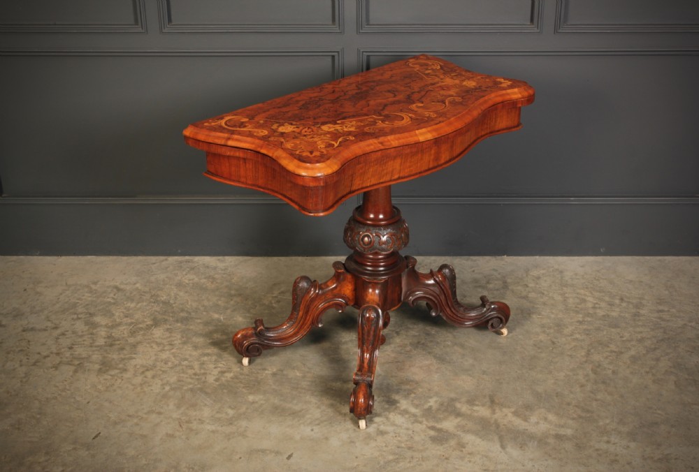 A Victorian figured walnut card table featuring floral marquetry inlay on a serpentine fold-over top, supported by a carved central stem and four cabriole legs on castors.