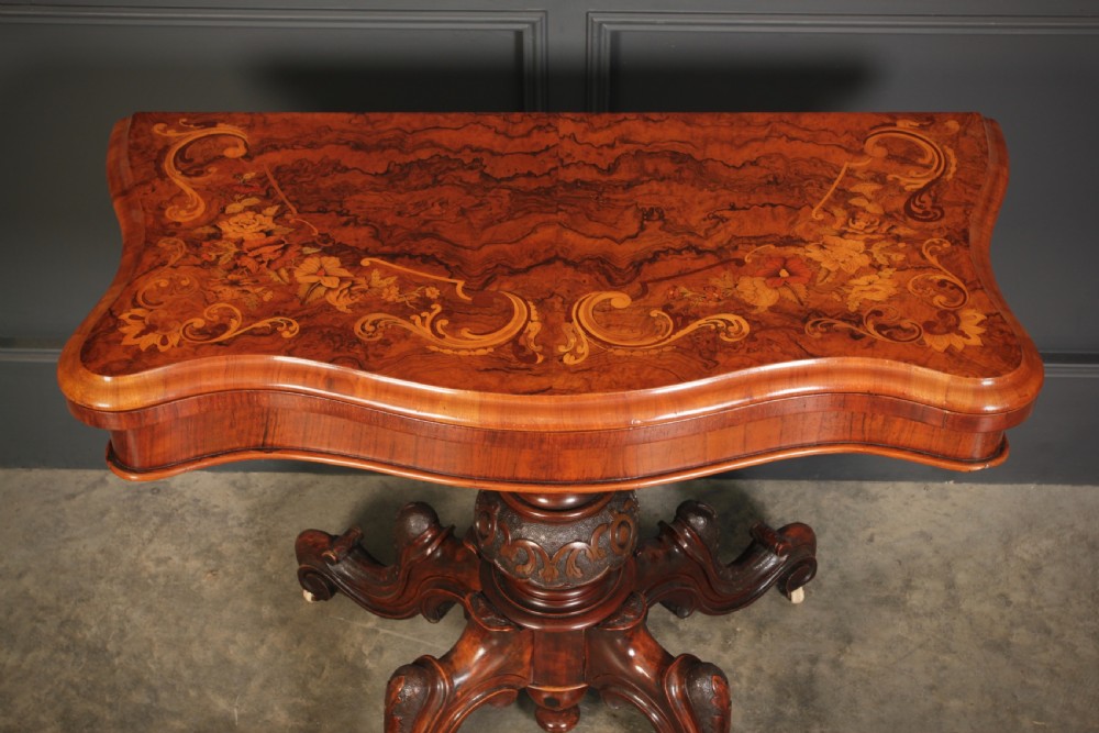 A Victorian figured walnut card table featuring floral marquetry inlay on a serpentine fold-over top, supported by a carved central stem and four cabriole legs on castors. View III