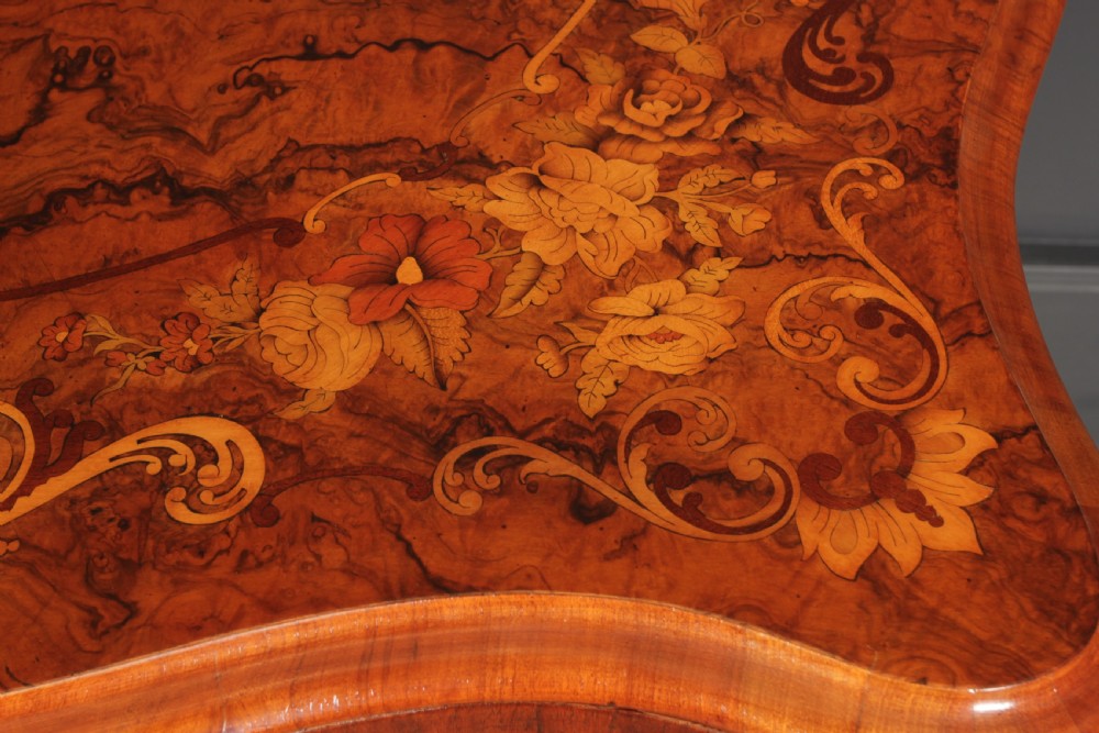 A Victorian figured walnut card table featuring floral marquetry inlay on a serpentine fold-over top, supported by a carved central stem and four cabriole legs on castors. View V