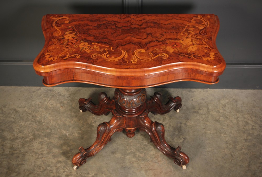 A Victorian figured walnut card table featuring floral marquetry inlay on a serpentine fold-over top, supported by a carved central stem and four cabriole legs on castors. View VII