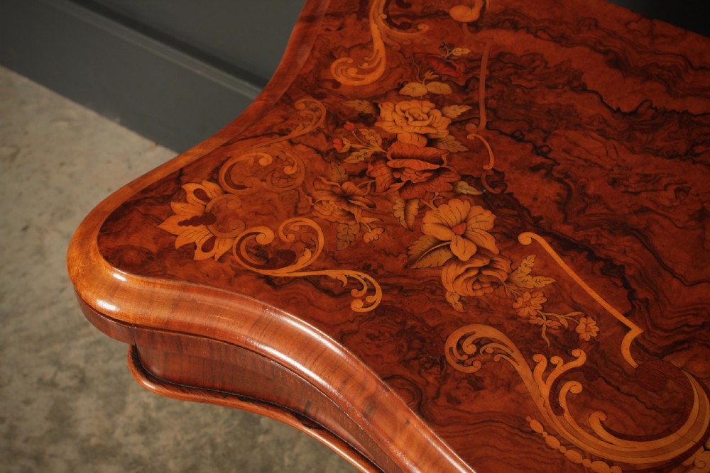 A Victorian figured walnut card table featuring floral marquetry inlay on a serpentine fold-over top, supported by a carved central stem and four cabriole legs on castors. View VIII