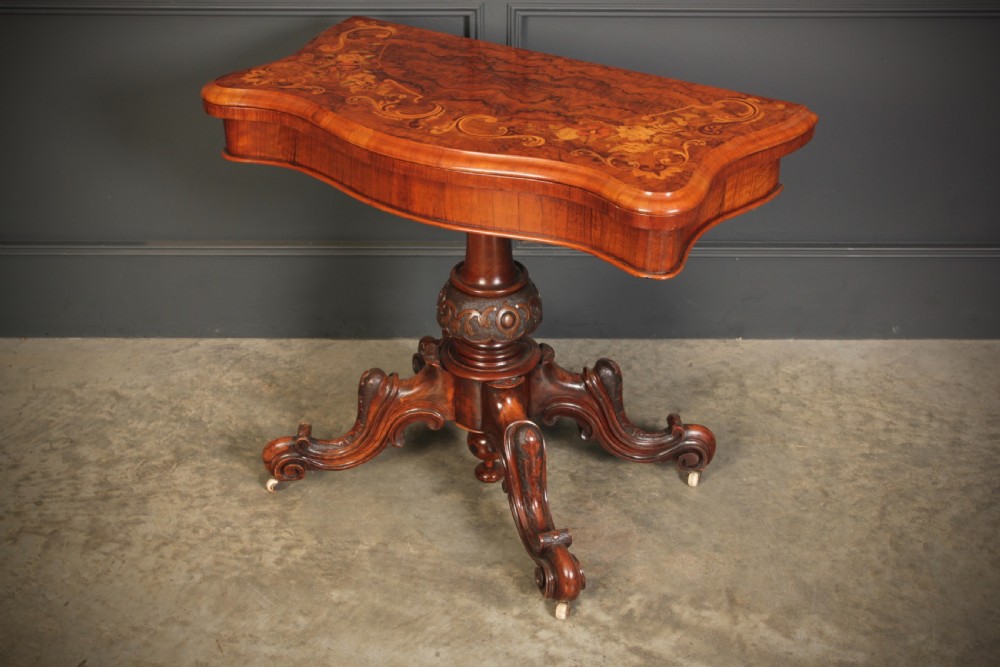 A Victorian figured walnut card table featuring floral marquetry inlay on a serpentine fold-over top, supported by a carved central stem and four cabriole legs on castors. View IX