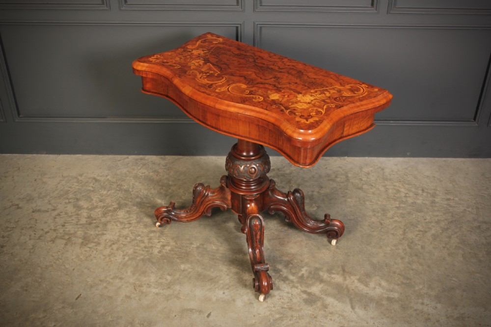 A Victorian figured walnut card table featuring floral marquetry inlay on a serpentine fold-over top, supported by a carved central stem and four cabriole legs on castors. View X