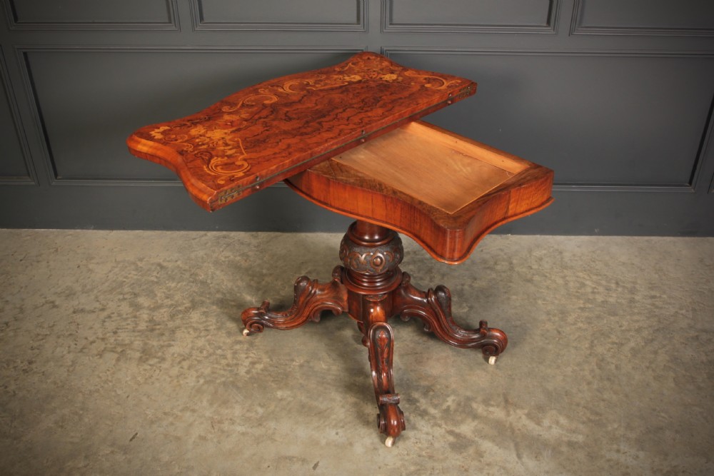 A Victorian figured walnut card table featuring floral marquetry inlay on a serpentine fold-over top, supported by a carved central stem and four cabriole legs on castors. View XI