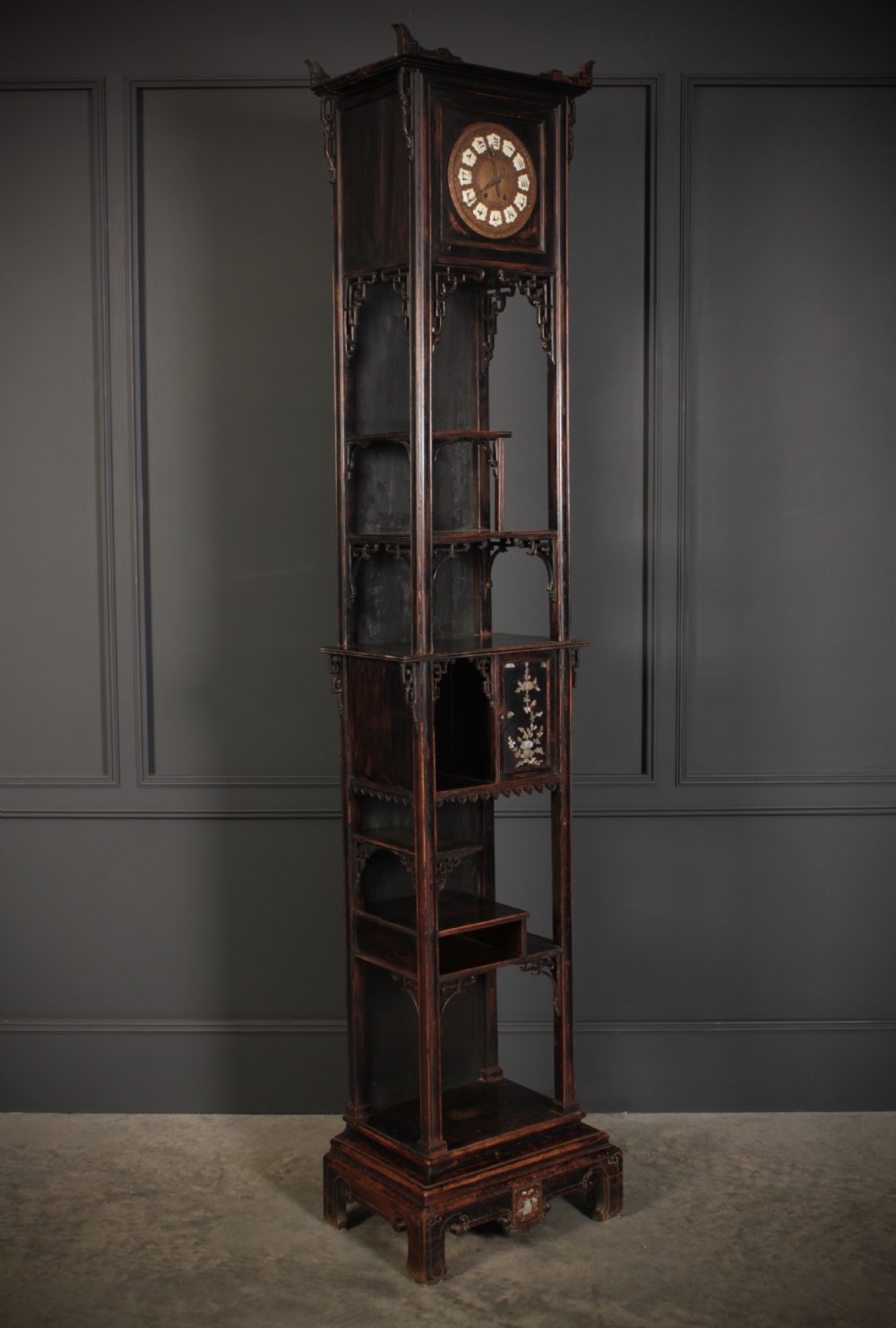 A tall, narrow antique Chinese dark wood floor clock featuring asymmetrical tiered display shelves, intricate carved fretwork, mother-of-pearl inlay details, and a pagoda-style bonnet top housing a circular clock face.