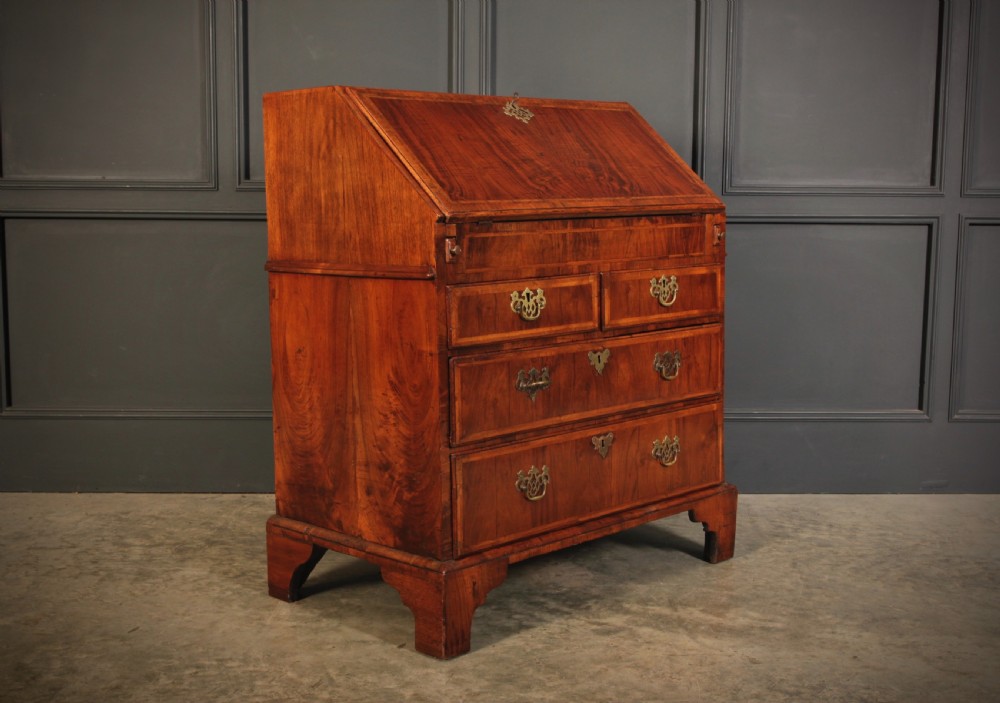 An eighteenth-century Queen Anne walnut slant-front bureau featuring brass drop handles, multiple drawers, and bracket feet.
