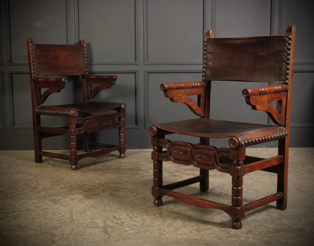 A pair of carved dark oak armchairs featuring stretched dark brown leather seats and backs fixed with brass metal studs.
