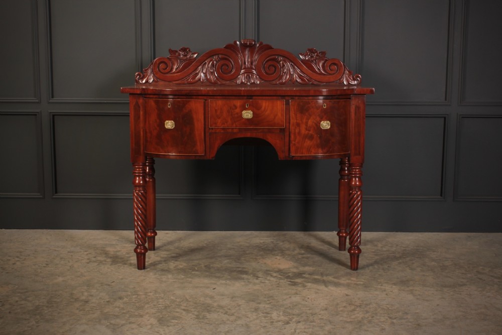 A William IV flame mahogany bow-front sideboard featuring a carved scroll backboard, flame veneer drawer fronts, brass keyhole escutcheons, and spiral-turned front legs. View V
