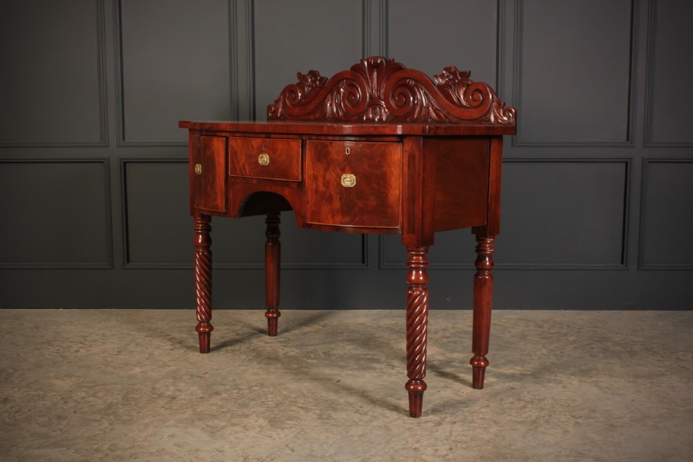 A William IV flame mahogany bow-front sideboard featuring a carved scroll backboard, flame veneer drawer fronts, brass keyhole escutcheons, and spiral-turned front legs. View II