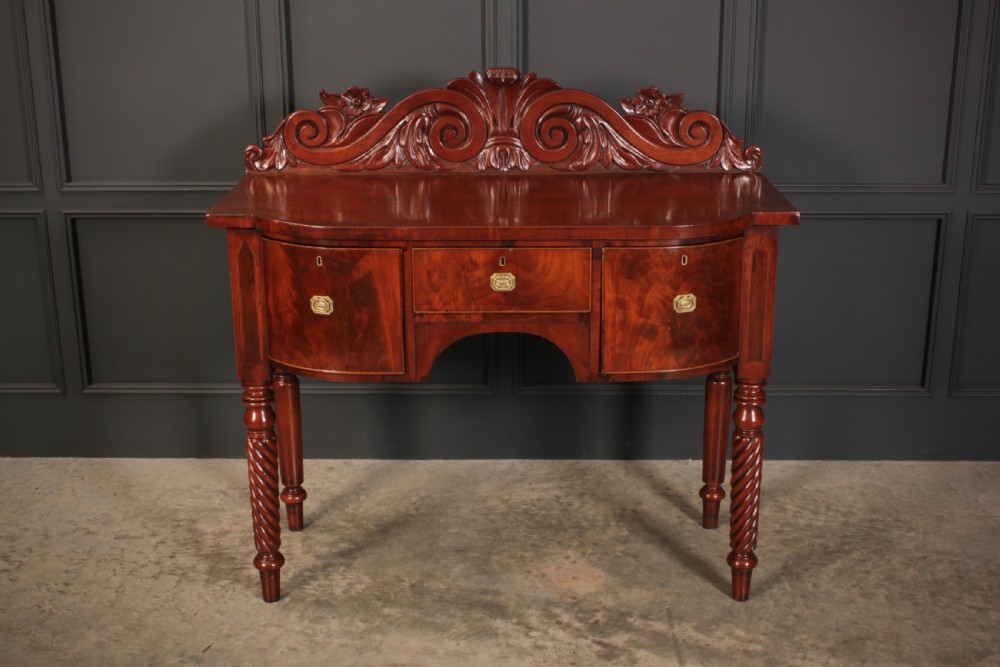 A William IV flame mahogany bow-front sideboard featuring a carved scroll backboard, flame veneer drawer fronts, brass keyhole escutcheons, and spiral-turned front legs. View III
