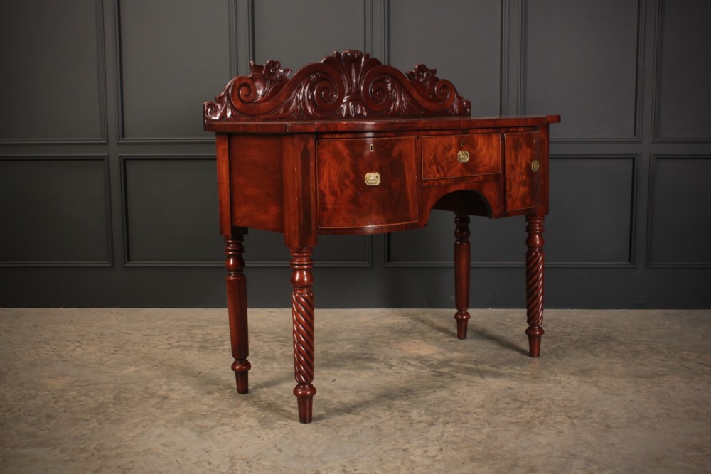 A William IV flame mahogany bow-front sideboard featuring a carved scroll backboard, flame veneer drawer fronts, brass keyhole escutcheons, and spiral-turned front legs.