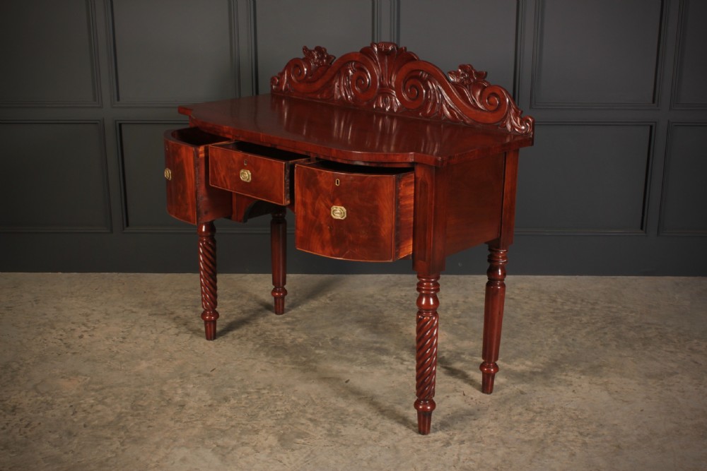 A William IV flame mahogany bow-front sideboard featuring a carved scroll backboard, flame veneer drawer fronts, brass keyhole escutcheons, and spiral-turned front legs. View VI