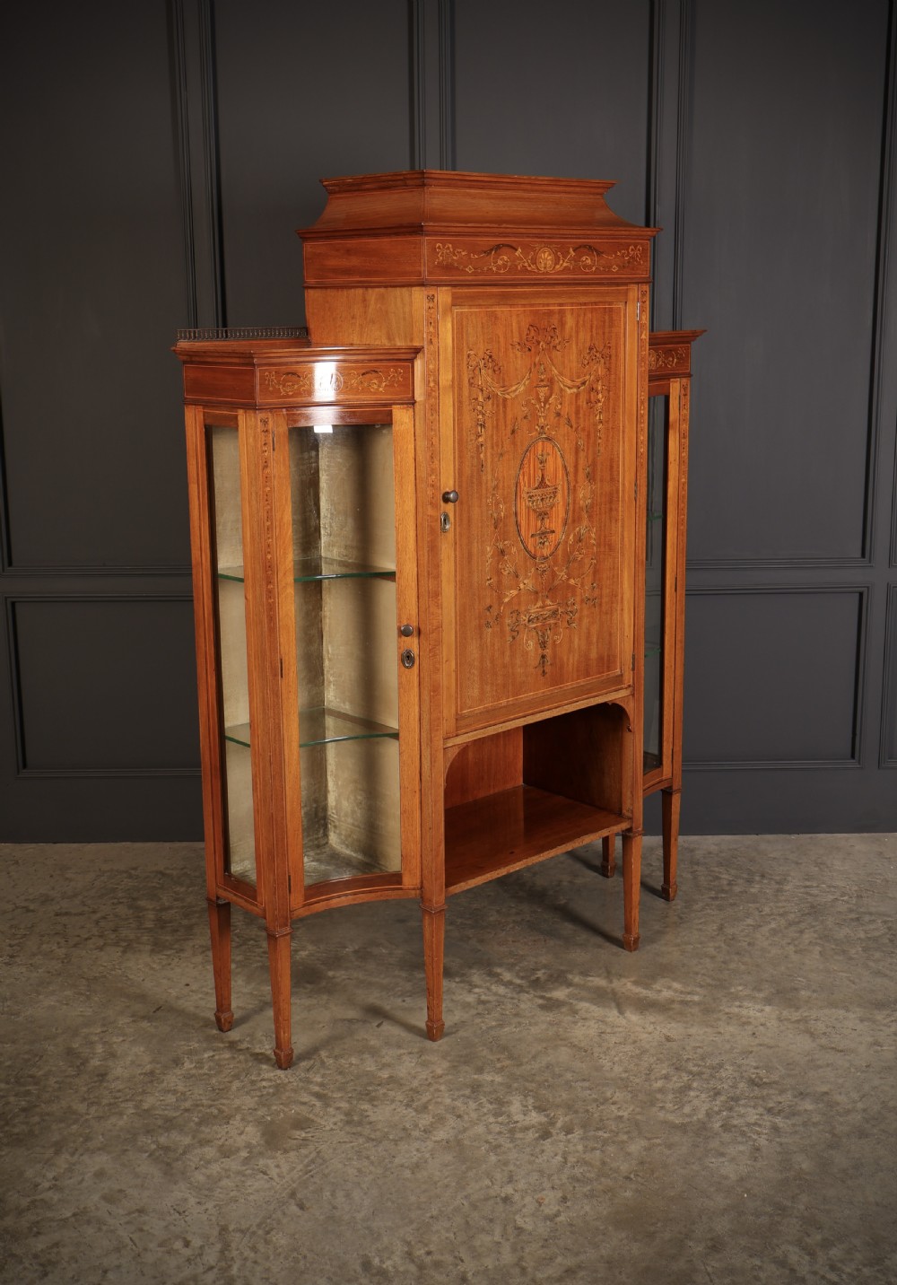 An Edwardian mahogany side cabinet with a central door featuring an urn marquetry inlay, flanked by two curved glazed display sections on tapered legs. View II