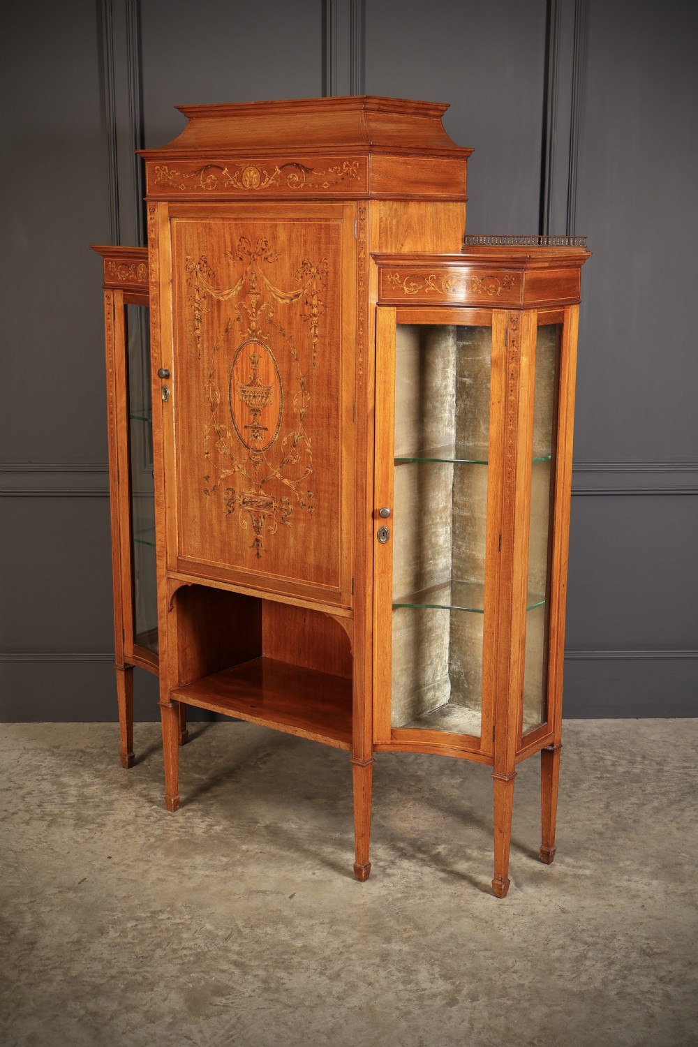 An Edwardian mahogany side cabinet with a central door featuring an urn marquetry inlay, flanked by two curved glazed display sections on tapered legs. View VIII
