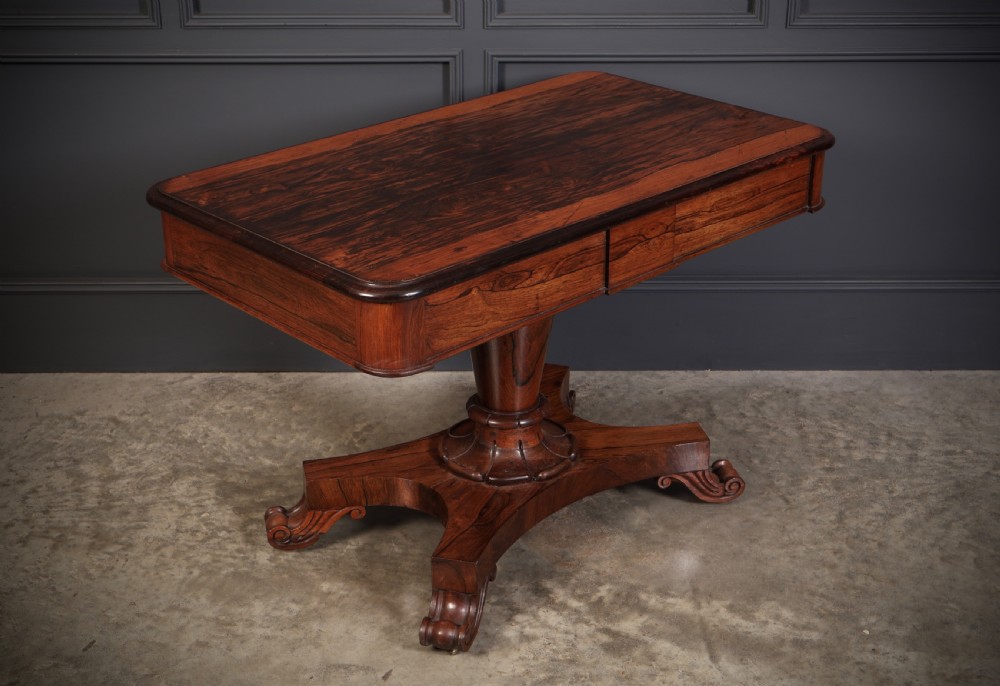 An antique rosewood library centre table featuring a rectangular figured top, two frieze drawers, a faceted pedestal column, and a quadform base with scroll feet.