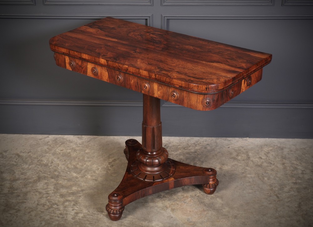 An antique Regency rosewood card table featuring a hinged rectangular top, rosette-decorated frieze, turned column, and triform pedestal base.