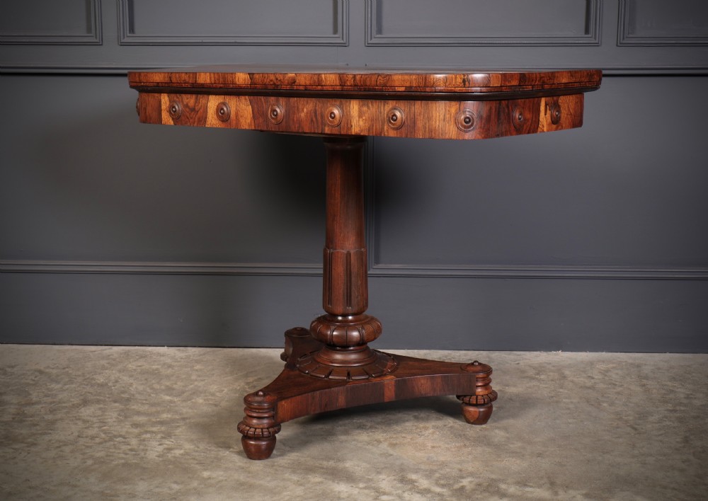 An antique Regency rosewood card table featuring a hinged rectangular top, rosette-decorated frieze, turned column, and triform pedestal base. View VI